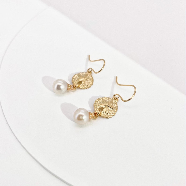 May include: A pair of gold earrings with a small pearl dangling from each. The earrings feature a textured, circular design with a star in the center.