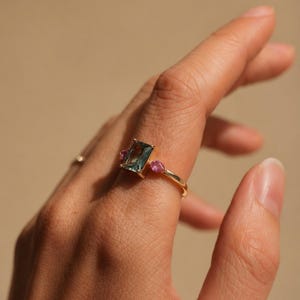 May include: A gold ring featuring a rectangular, teal-colored gemstone flanked by two smaller, pink gemstones. The ring is set on a finger, showcasing the design and color contrast against the skin.