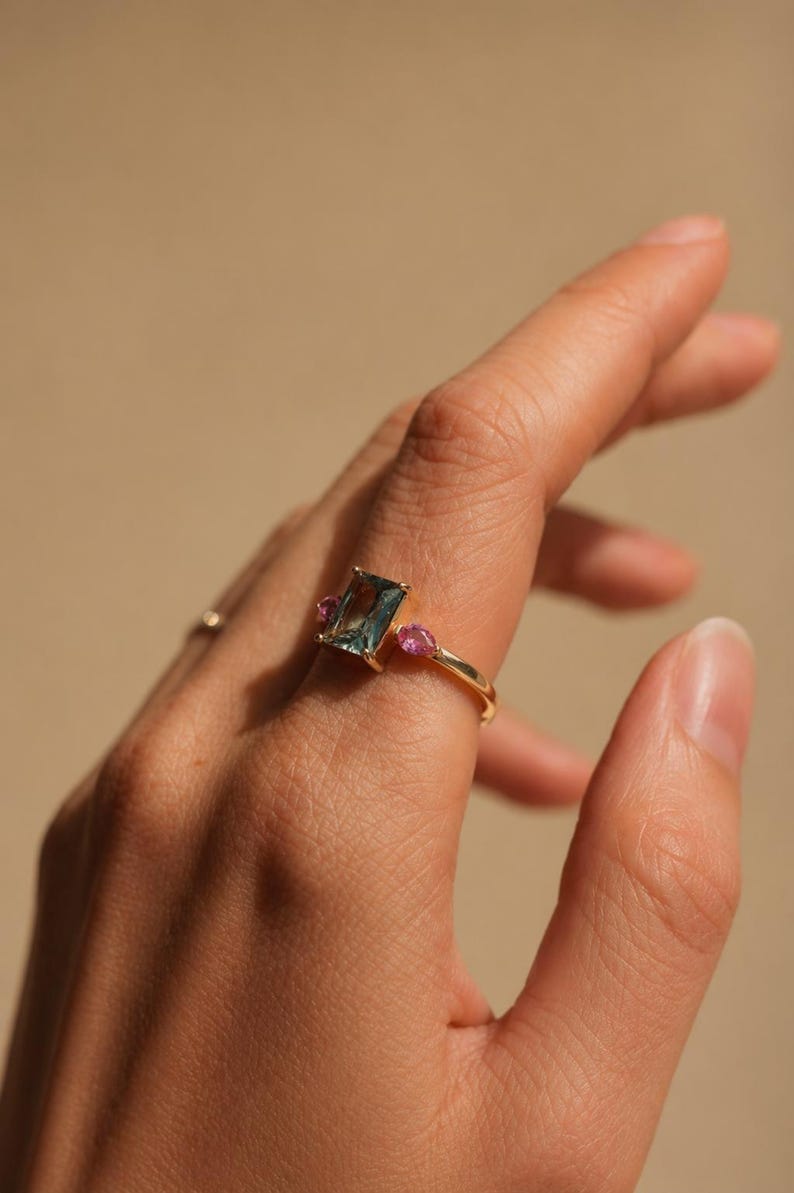 May include: A gold ring featuring a rectangular, teal-colored gemstone flanked by two smaller, pink gemstones. The ring is set on a finger, showcasing the design and color contrast against the skin.