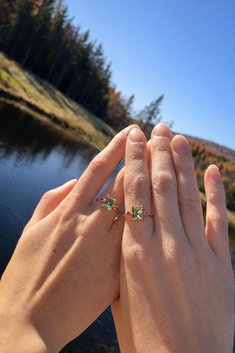 May include: Two hands are shown, each wearing a gold ring. The rings feature a square, light green gemstone flanked by smaller pink gemstones. The background includes a lake and trees under a blue sky.