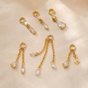 May include: A collection of six gold-toned metal jewelry charms featuring clear, faceted crystals. The charms include various designs such as single drops, multi-chain dangles, and a star-shaped accent, all attached to small circular jump rings.