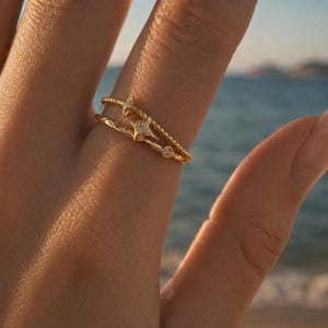 May include: Two gold rings, one with a star design and a small diamond, the other with a textured band and a beaded band with a small diamond. The rings are worn on a finger, with a blurred ocean background.