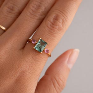 May include: A gold ring featuring a rectangular, light blue gemstone flanked by two smaller, pink gemstones. The ring is set on a hand, with a simple gold band visible in the background. The ring's design is elegant and modern.
