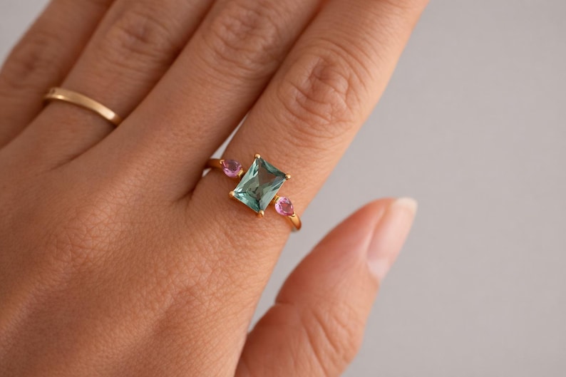 May include: A gold ring featuring a rectangular, light blue gemstone flanked by two smaller, pink gemstones. The ring is set on a hand, with a simple gold band visible in the background. The ring's design is elegant and modern.