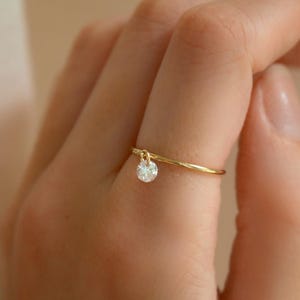 Peut inclure: Une délicate bague en or avec un petit diamant rond qui pend. La bague est portée à un doigt.