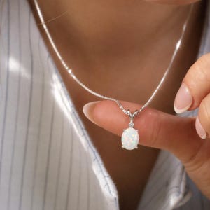 Puede incluir: Un collar de plata con un pequeño colgante ovalado de ópalo blanco. El colgante está sostenido por una mano, y el collar está contra una camisa a rayas.
