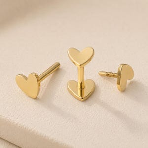 May include: Three gold-colored heart-shaped earrings. One earring has a post, another has a flat base and post, and the third has a threaded post. The earrings are displayed on a light-colored surface.