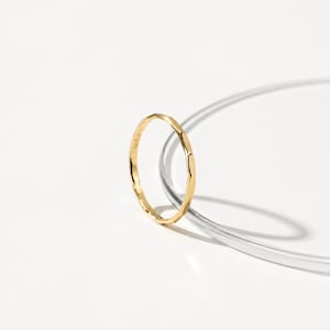 Peut inclure: Une fine bague en or avec un design facetté. La bague est mince et présente une surface texturée, captant la lumière. Elle est posée sur un fond blanc avec un élément incurvé et transparent.
