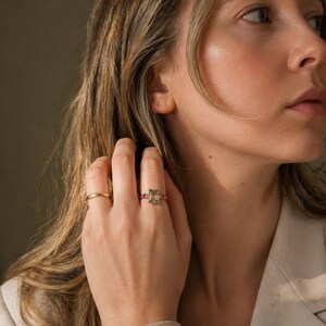 May include: A gold ring with a rectangular, light green gemstone and small pink stones. A second, simple gold band ring is also visible. The rings are on a hand, with a neutral background.