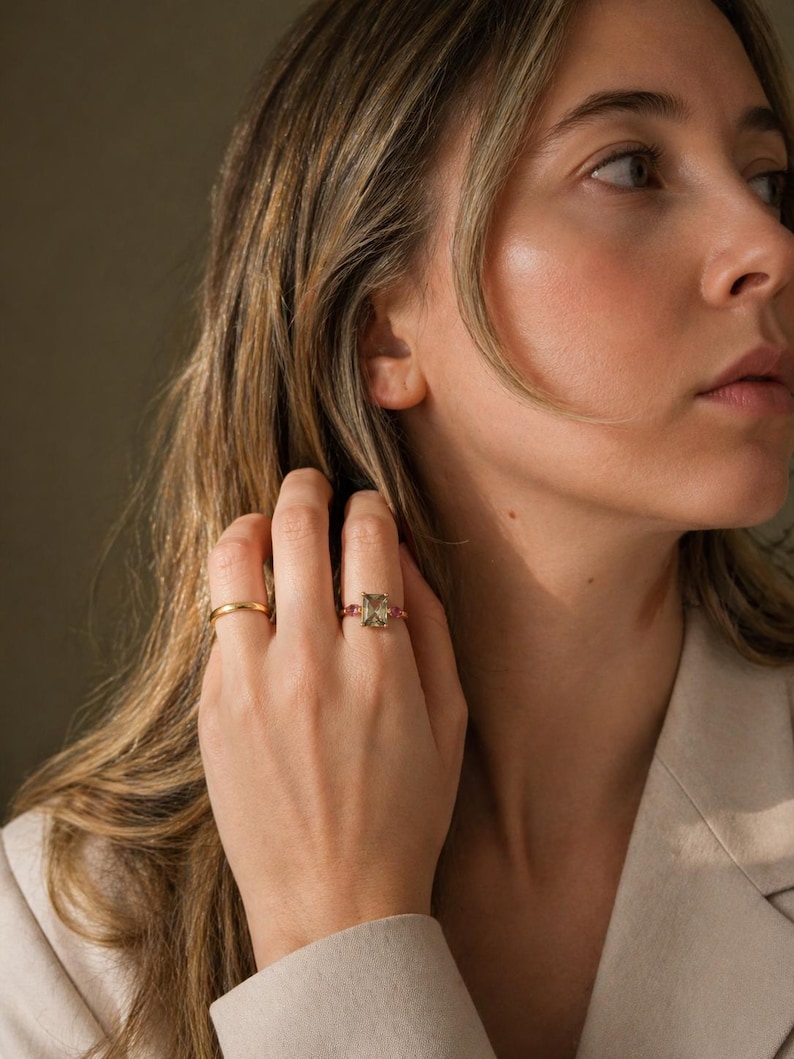 May include: A gold ring with a rectangular, light green gemstone and small pink stones. A second, simple gold band ring is also visible. The rings are on a hand, with a neutral background.