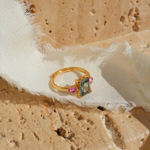 May include: A gold ring with a rectangular, bluish-green center stone and two pink side stones. The ring is set on a white fabric against a textured, beige surface. The ring's design is elegant and modern.