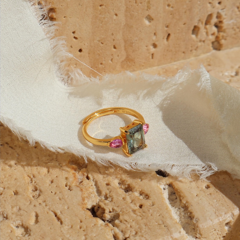 May include: A gold ring with a rectangular, bluish-green center stone and two pink side stones. The ring is set on a white fabric against a textured, beige surface. The ring's design is elegant and modern.