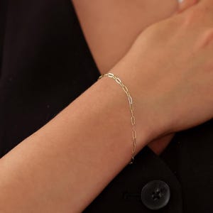 May include: A delicate gold chain bracelet with an elongated oval link design. The bracelet is worn on a wrist, showcasing its simple yet elegant style. The bracelet's links are approximately 0.5 inches in length.