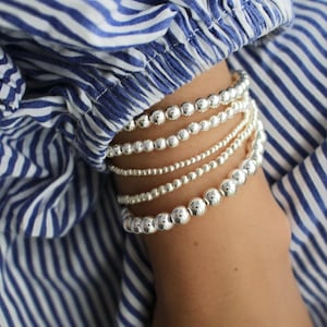 May include: A set of five stacked stretch bracelets featuring polished silver-toned spherical beads of varying sizes. The bracelets are arranged in a row, showcasing a reflective metallic finish and a uniform circular shape on each individual strand.