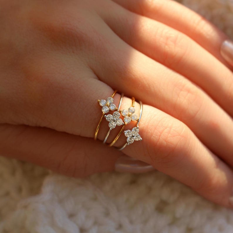 May include: Three gold rings with delicate flower-shaped designs set with clear sparkling stones. The rings are stacked on a finger.