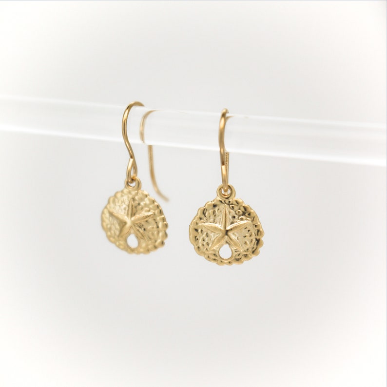 May include: Gold dangle earrings with a sand dollar design. The sand dollar has a five-pointed star in the center.