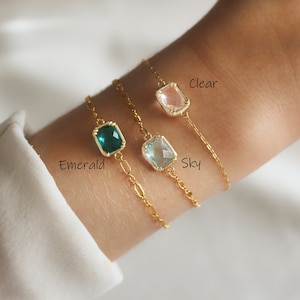 May include: Three gold chain bracelets with different coloured square-shaped glass charms. The charms are labelled "Clear", "Sky", and "Emerald".