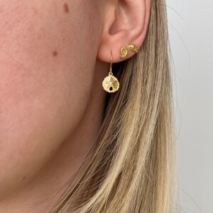 May include: Gold dangle earrings featuring a seashell design. The earrings are shown on a model with blonde hair.