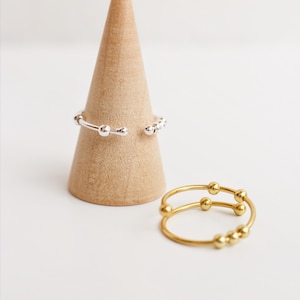May include: Two rings, one silver and one gold, are shown against a light backdrop. The silver ring has an open design with small ball details. The gold ring has a double band with ball accents. A wooden cone is in the background.