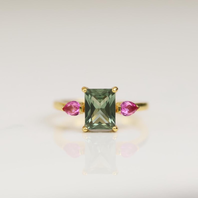 May include: A gold ring featuring a rectangular, olive-green gemstone flanked by two pink teardrop-shaped stones. The ring is set against a white background, highlighting the jewelry's design and color contrast.