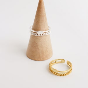May include: Two silver rings with a chain design, one with a small oval shape, and a gold ring with a chain design, all displayed on a wooden ring holder.