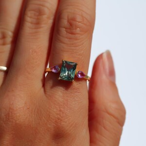 May include: A gold ring with a rectangular, teal-colored center stone and two smaller, pink teardrop-shaped stones. A simple silver band ring is also visible. The rings are on a person's hand.