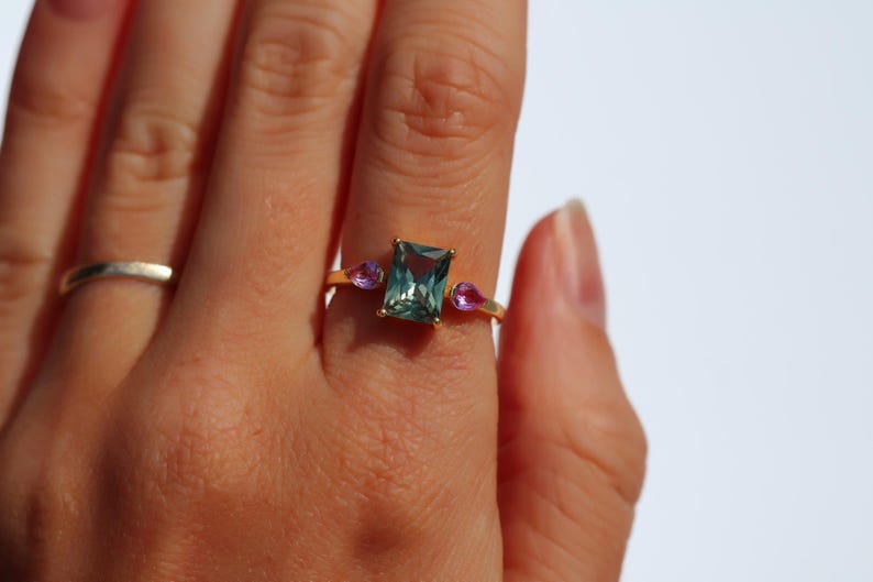 May include: A gold ring with a rectangular, teal-colored center stone and two smaller, pink teardrop-shaped stones. A simple silver band ring is also visible. The rings are on a person's hand.