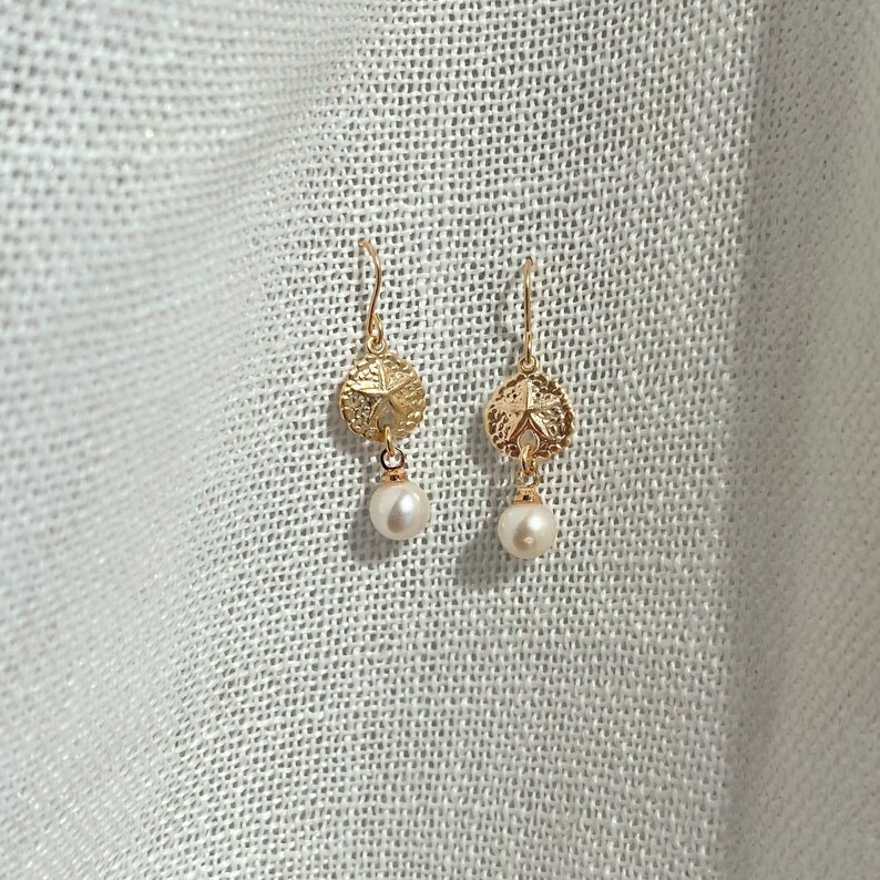 May include: Gold earrings with a sand dollar charm and a pearl drop.