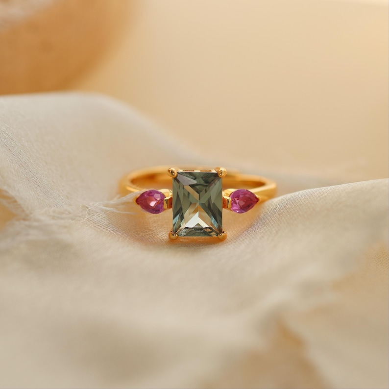 May include: A gold ring featuring a rectangular, light green gemstone as the centerpiece, flanked by two smaller, teardrop-shaped pink gemstones. The ring is set against a soft, cream-colored fabric.