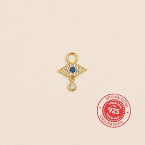 May include: A gold-coloured charm with a diamond-shaped design, featuring a blue centre stone and surrounding clear stones. A smaller, dangling clear stone hangs below. The charm has a loop at the top for attachment.