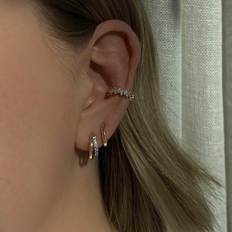 K&ouml;nnte beinhalten: Nahaufnahme eines Ohrs mit drei goldenen Ohrringen. Der obere Ohrring ist ein funkelnder, mit Steinen besetzter Ohrclip. Die beiden unteren Ohrringe sind d&uuml;nne, goldene Creolen.