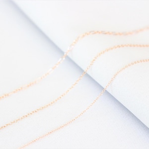 May include: Three delicate gold chains lay on a white surface. The chains are arranged in a staggered pattern, with the top chain being the longest and the bottom chain being the shortest.