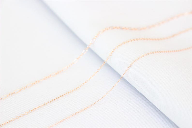 May include: Three delicate gold chains lay on a white surface. The chains are arranged in a staggered pattern, with the top chain being the longest and the bottom chain being the shortest.