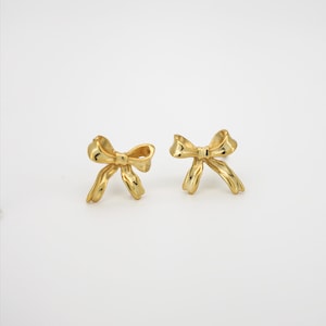 May include: A pair of gold-toned stud earrings shaped like small bows. Each earring features a polished, reflective metallic surface with a central knot and two flowing ribbon ends, designed with a smooth, curved silhouette and a classic bow form.