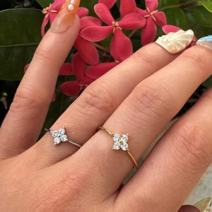 May include: Two dainty rings with a flower design featuring small clear stones. The ring on the left is silver and the ring on the right is gold.