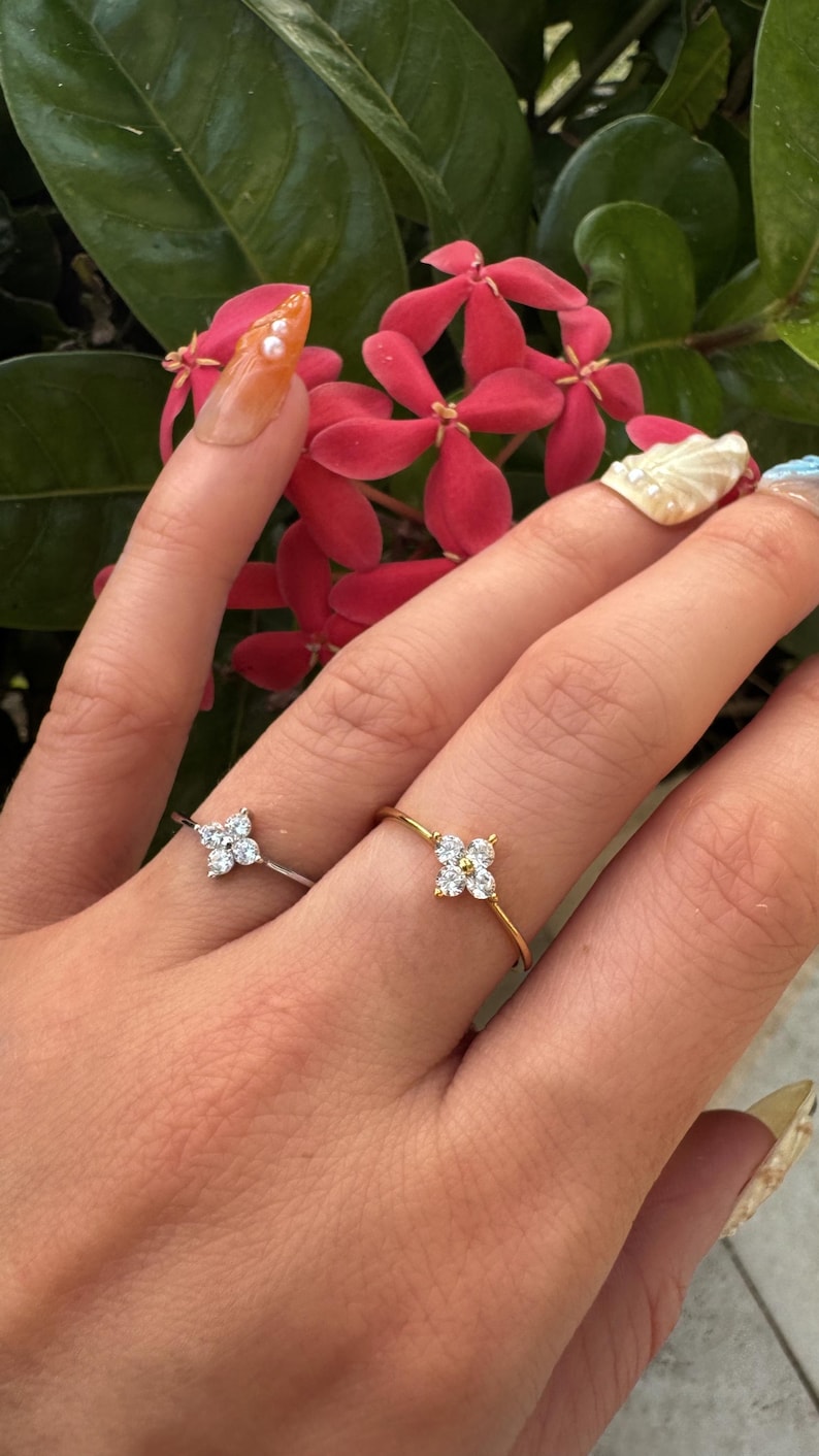 May include: Two dainty rings with a flower design featuring small clear stones. The ring on the left is silver and the ring on the right is gold.