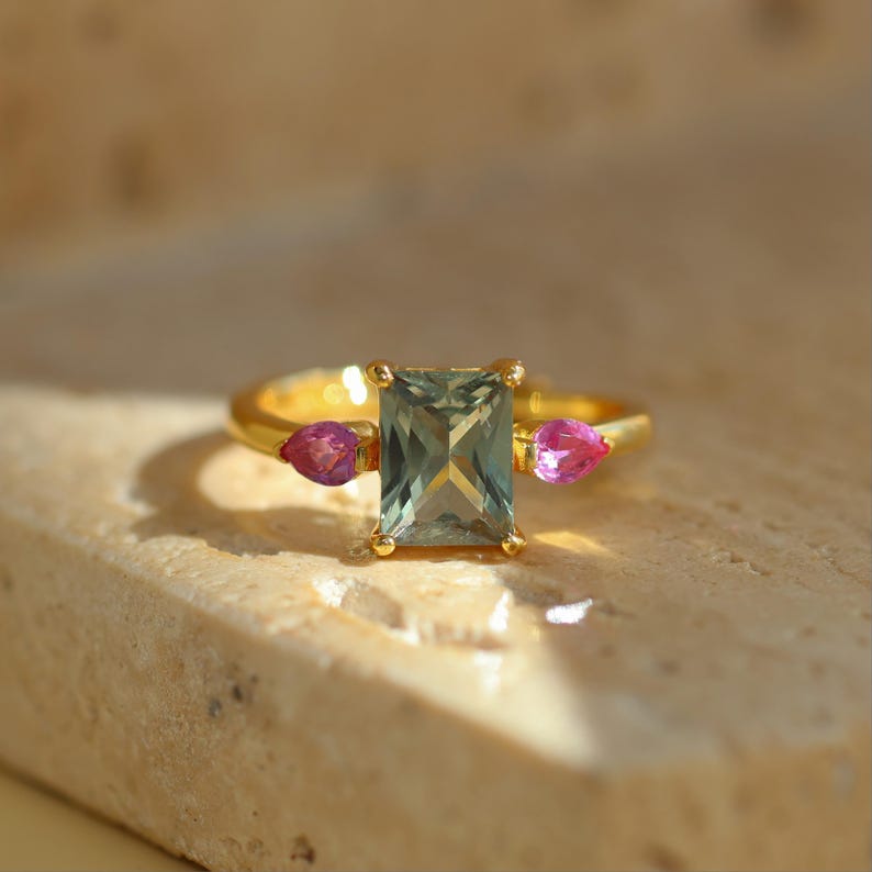 May include: A gold ring featuring a rectangular, light green gemstone as the centerpiece, flanked by two smaller, pink teardrop-shaped stones. The ring is set on a neutral-toned surface, showcasing the jewelry's design and color contrast.