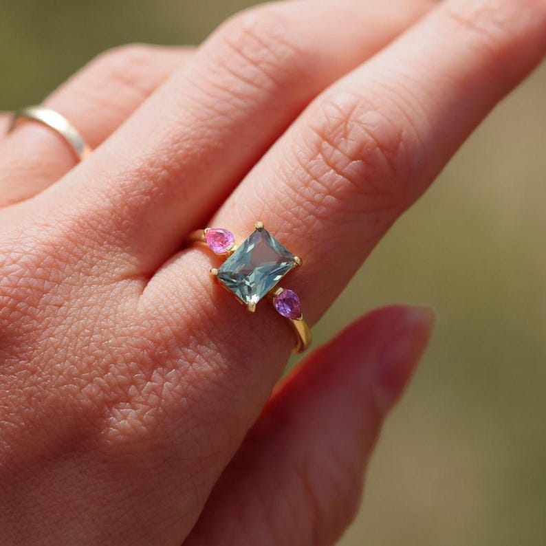 May include: A gold ring featuring a rectangular, light blue gemstone flanked by two smaller, teardrop-shaped pink and purple gemstones. The ring is set on a finger, showcasing the jewelry's design and color contrast.