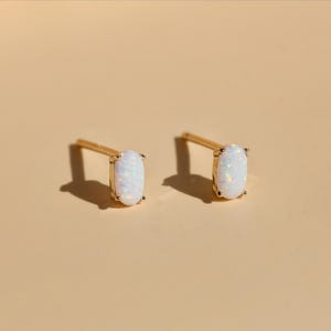 May include: A pair of gold stud earrings, each featuring an oval-shaped, iridescent white opal stone. The earrings are set in a gold bezel setting and have a post backing. The background is a solid, light beige color.