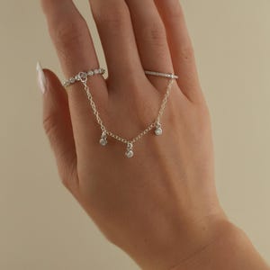 Könnte beinhalten: Eine silberne Handkette mit drei baumelnden Anhängern ist mit zwei Fingerringen verbunden. Die Ringe und Anhänger sind mit kleinen, klaren Edelsteinen besetzt. Der Schmuck wird auf einer Hand vor neutralem Hintergrund präsentiert.