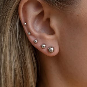 May include: A close-up shot of an ear adorned with a series of silver ball stud earrings. The earrings are graded in size, from small to large. The ear is positioned against a background of blonde hair, creating a simple yet elegant aesthetic.