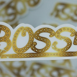 May include: Gold sticker with the word "Bossy" in a decorative font with a border of small circles. The sticker is on a white background.