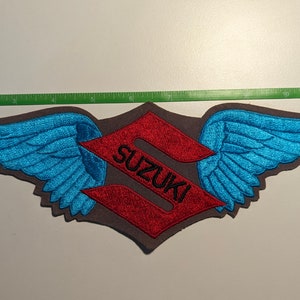 May include: A red and black embroidered patch with the word "SUZUKI" and wings on either side. The wings are blue and have a feathered design.