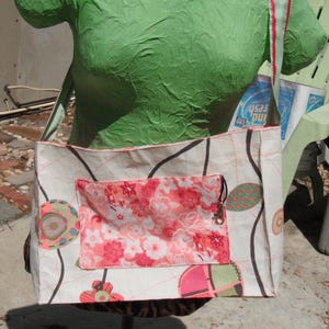 May include: A white canvas tote bag with a pink floral pocket and a black strap. The bag has a colorful design with circles and swirls. The bag is being held by a green mannequin.
