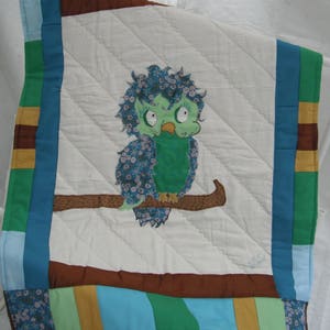 May include: A handmade quilt featuring a whimsical owl design. The owl, in shades of green and blue, is perched on a brown branch against a white background. The quilt's border includes various colorful fabric strips.