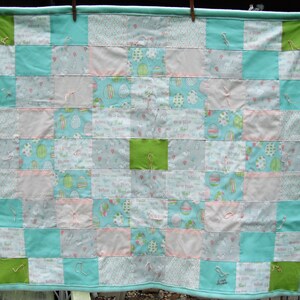 May include: A patchwork quilt with a light blue border and backing. The quilt features squares of various patterns, including Easter eggs, text, and animals, in shades of blue, green, and pink. The quilt is hung on a washing line.