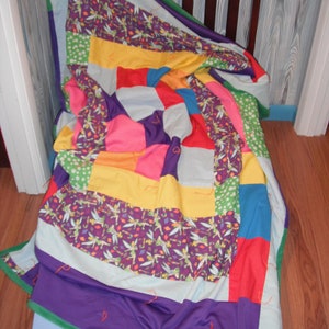 May include: A colorful patchwork quilt with a variety of fabrics and patterns. The quilt features squares of purple, pink, yellow, red, and blue, with a section of purple fabric with a fairy print. The quilt is draped over a light blue surface.
