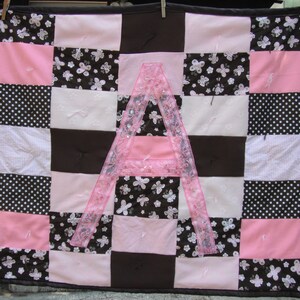 May include: A pink and brown patchwork quilt with a large pink letter A in the center. The quilt is made of various fabrics with different patterns, including polka dots, butterflies, and stripes.