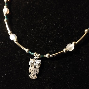 May include: A silver necklace with white beads and a silver owl charm. The necklace is on a black background.