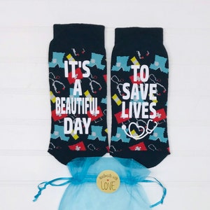 May include: A pair of black socks with a colourful medical theme print. One sock reads "It's a beautiful day" and the other reads "To save lives". The socks are packaged in a light blue organza bag with a wooden tag that reads "Handmade with love".